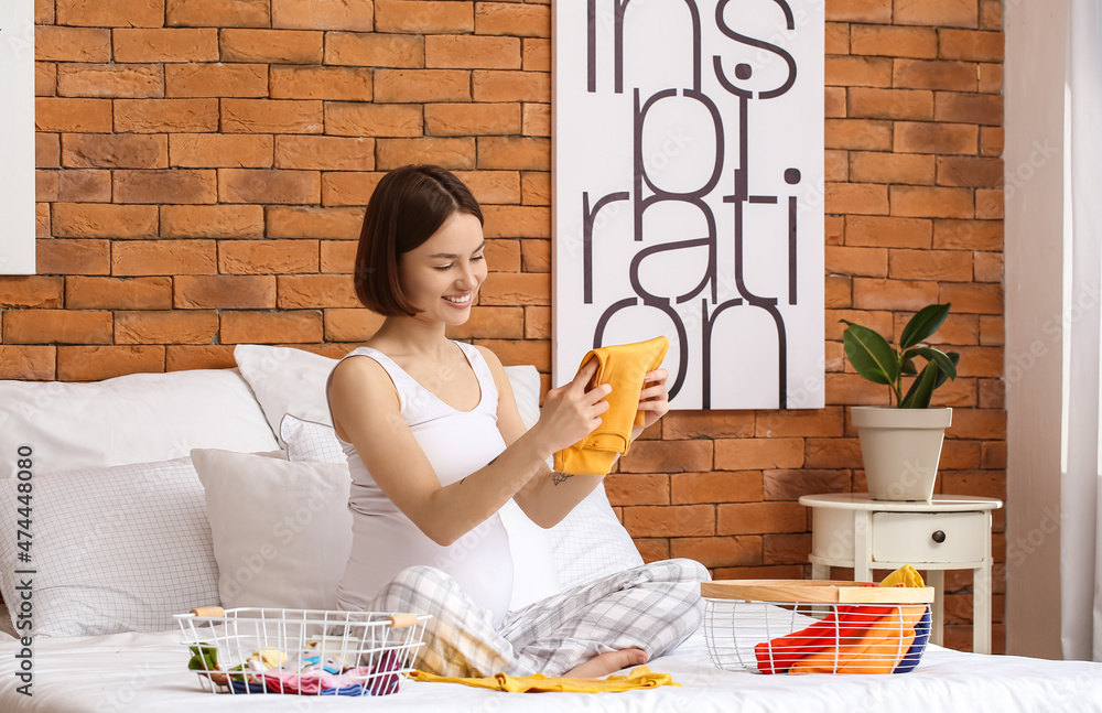 Young pregnant woman putting baby clothes into basket in bedroom