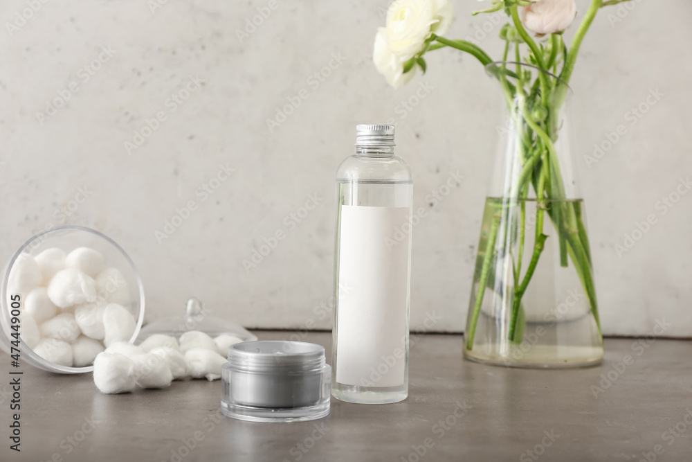 Bottle with cosmetics, jar with face cream and vase with ranunculus flowers on grey table