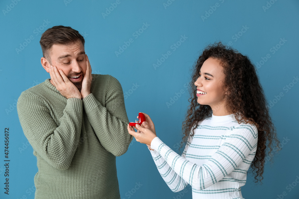 Young woman proposing to her happy boyfriend on color background