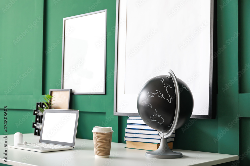 Modern workplace with laptop and blank frames hanging on green wall