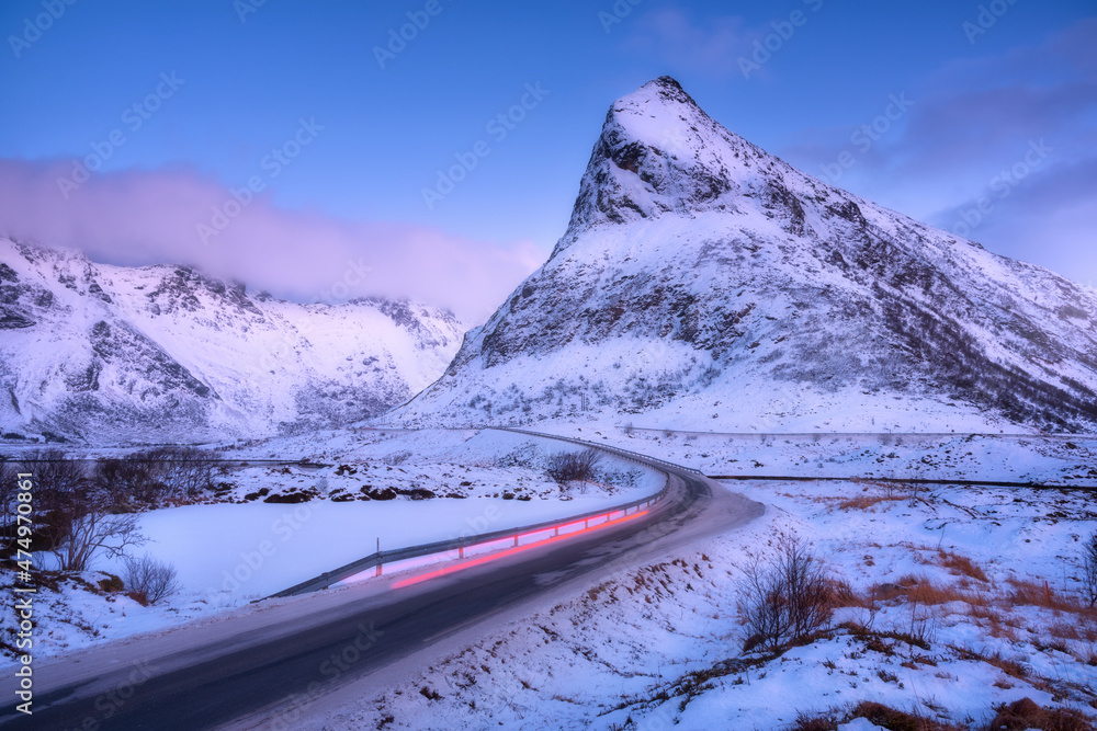 冬天黄昏时，道路上的雪山和红色模糊的汽车前灯。挪威罗弗敦群岛