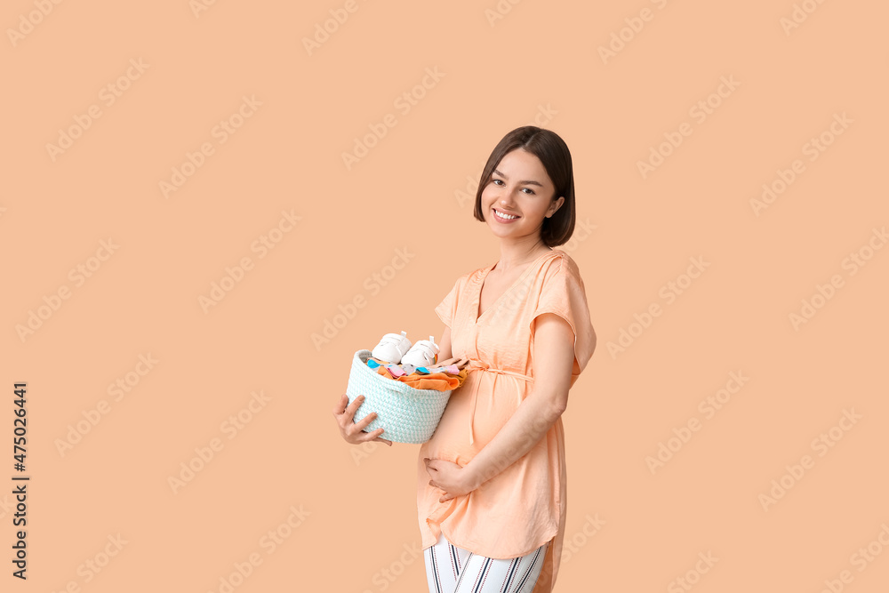 Young pregnant woman holding basket with baby clothes on beige background