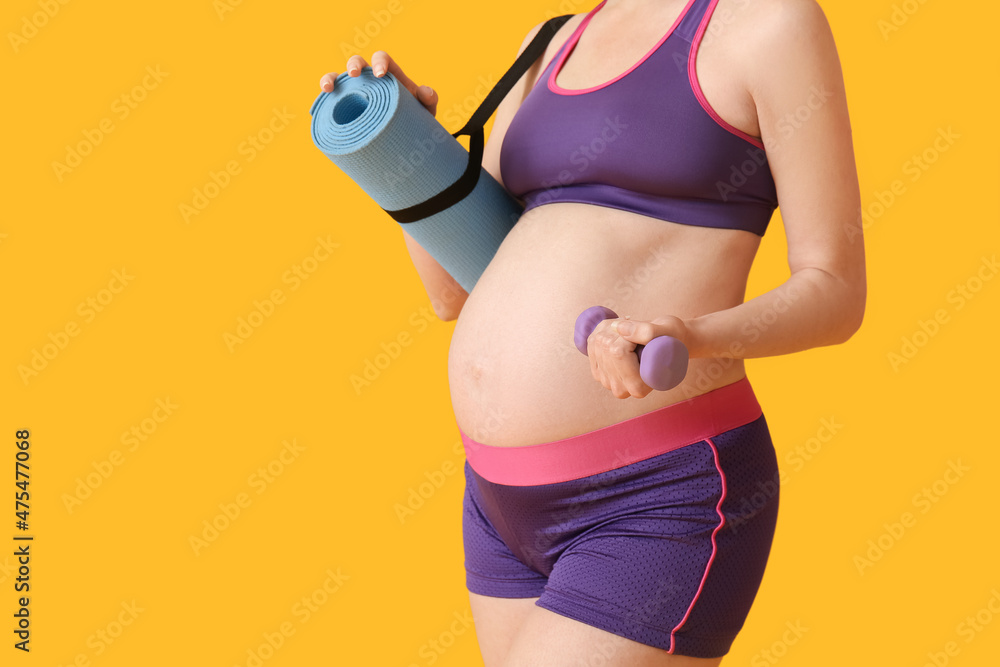 Young pregnant woman with dumbbell and yoga mat on yellow background