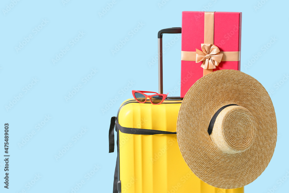 Yellow suitcase with sunglasses, Christmas gift box and hat on blue background