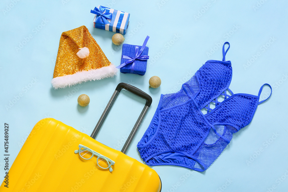 Yellow suitcase with Christmas gift boxes, balls, Santa hat and swimsuit on blue background