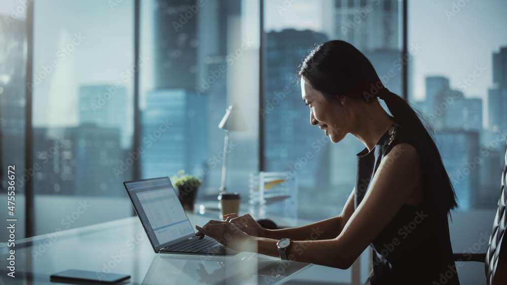 Successful Businesswoman in Stylish Dress Sitting at a Desk in Modern Office, Using Laptop Computer,