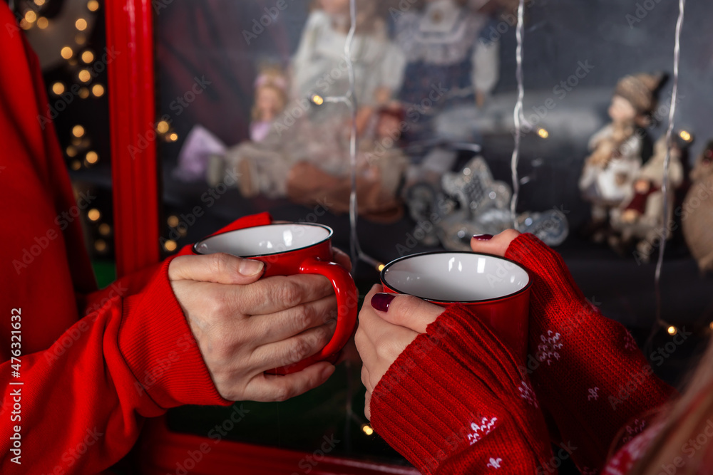 Girls hands in warm sweaters holding red cups. close up. background with Chistmas decoration