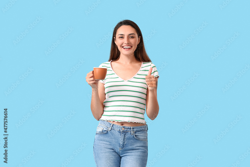 Beautiful woman with cup of coffee showing thumb-up on color background