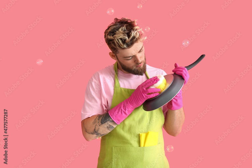 Handsome young man with sponge cleaning frying pan on pink background