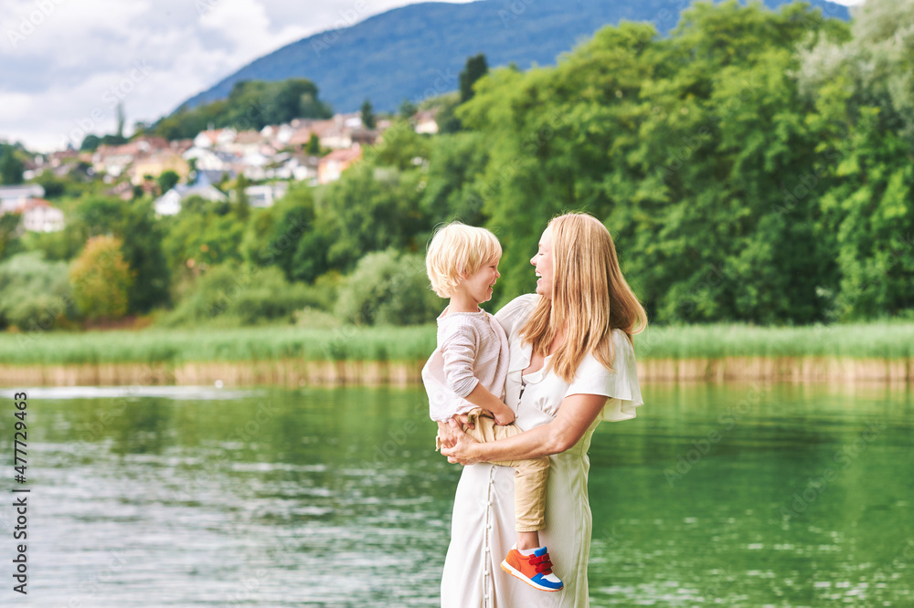 Outdoor portrait of happy young mother with adorable preschooler son, enjoying nice day next to lake