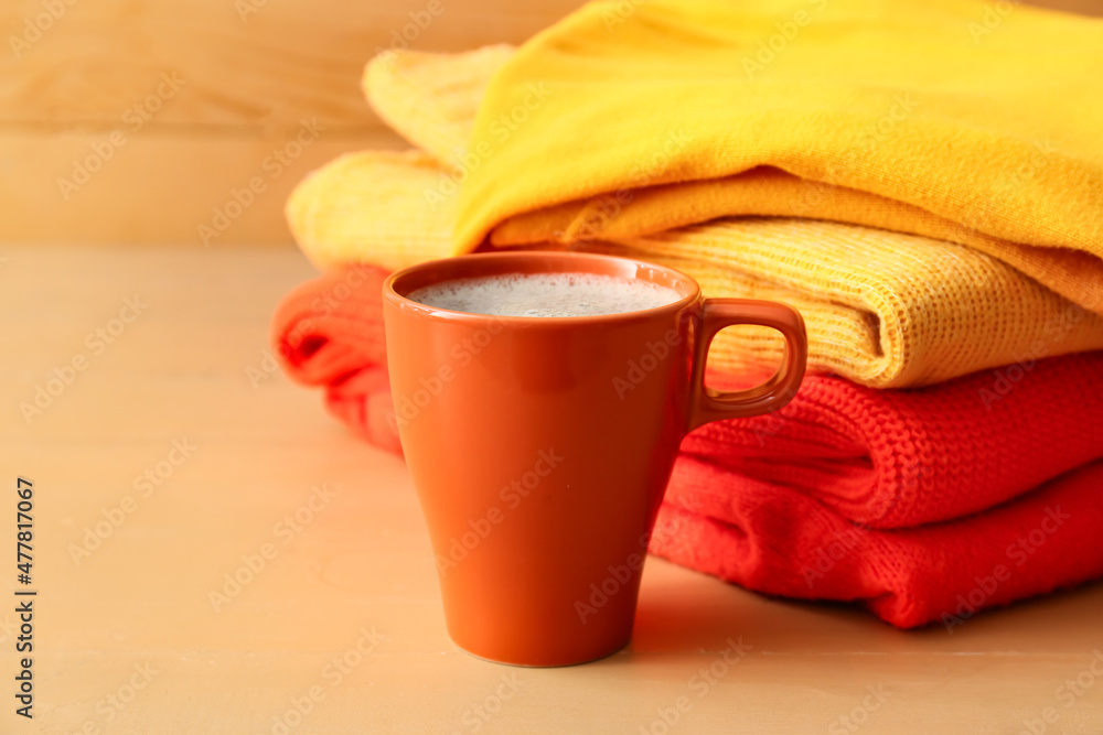 Stack of different cozy sweaters and cup of coffee on wooden background