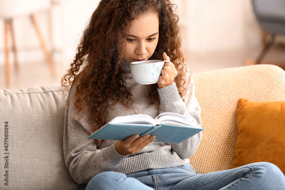 Young African-American woman drinking tea while reading book on sofa at home