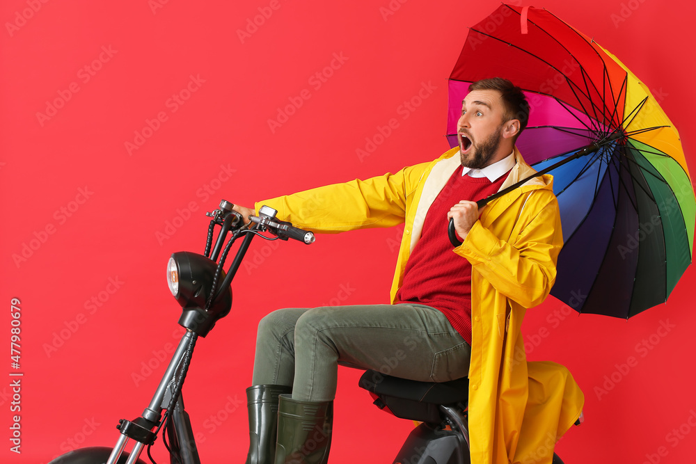 Surprised man in raincoat and with umbrella riding bike on color background