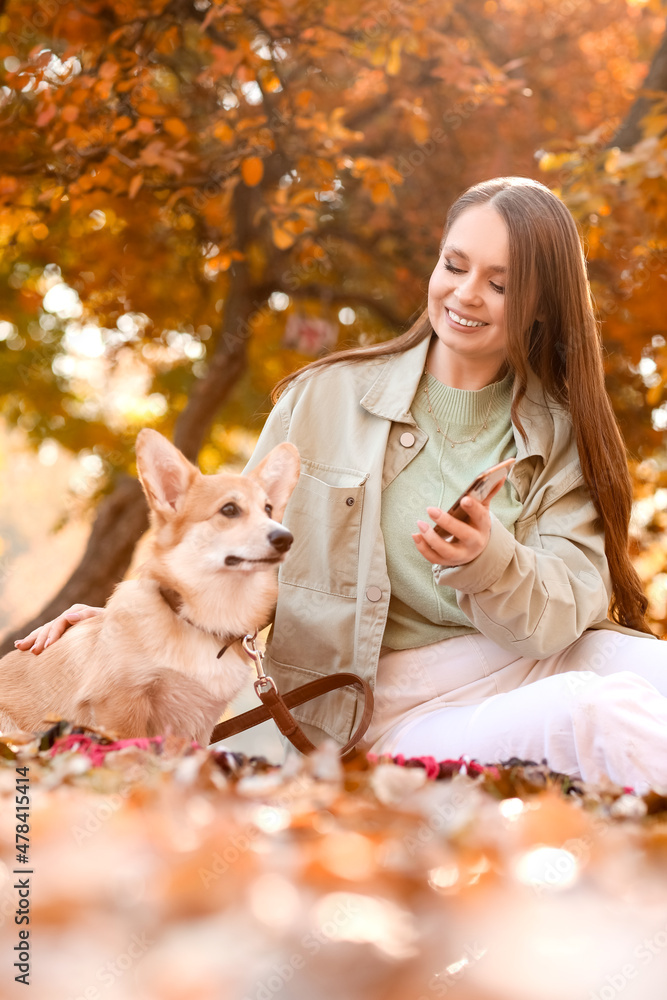 带着手机的年轻女子和可爱的柯基犬坐在秋天公园的格子上