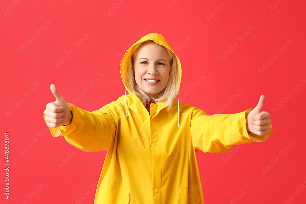 Beautiful mature woman in raincoat showing thumb-up on color background