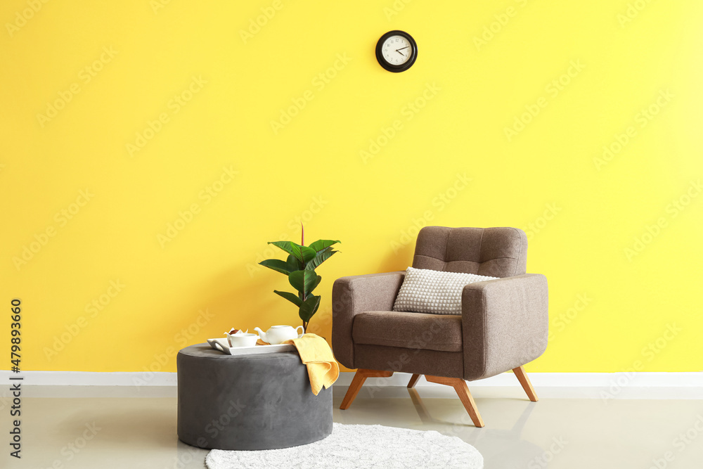 Pouf with breakfast, houseplant and soft armchair near yellow wall