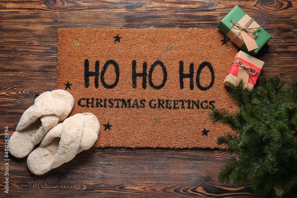 Door mat with Christmas greeting, slippers, gifts and fir tree on dark wooden floor