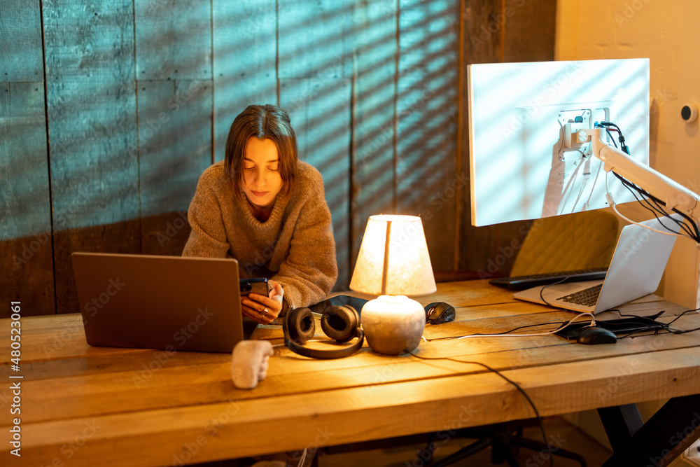 Young focused woman using smart phone while sitting at cozy home office during the nighttime. Concep