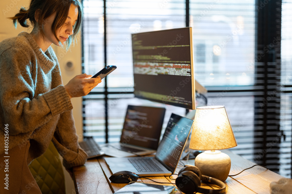 Young woman using phone while sitting at workplace with laptop and desktop computers at cozy home of