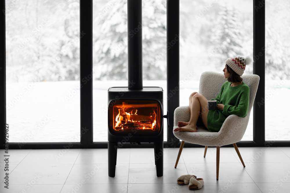 Woman sitting with cup on chair by the fireplace at modern house on nature during winter time. Conce