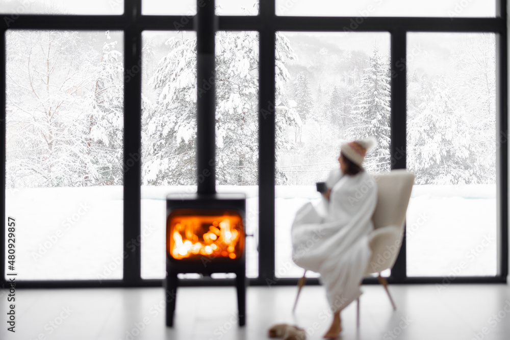 Woman covered with blanket enjoys winter time at home with burning fireplace, looking outside the wi