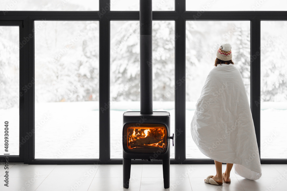 Woman enjoys winter time at home with burning fireplace and panoramic window, looking outside on sno