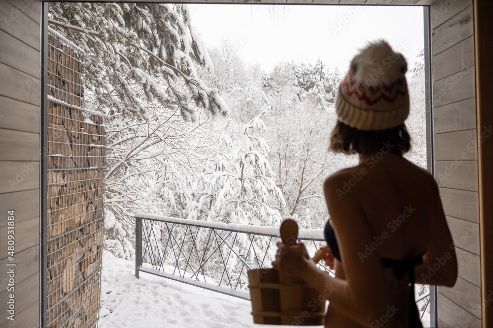 Young woman standing with backet in sauna with beautiful view on snowy forest. Concept of winter SPA