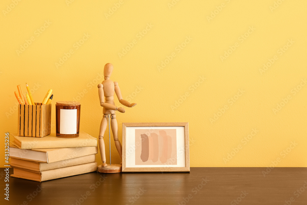 Stack of books, stationery holder and wooden mannequin doll on table near color wall