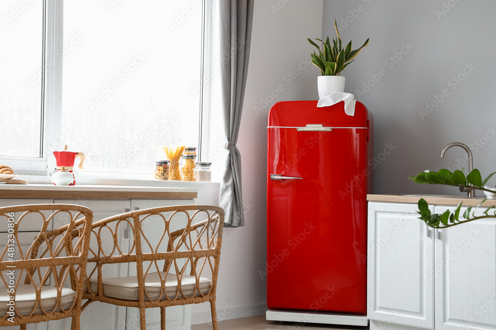 Interior of light modern kitchen with red fridge, white counters and wicker chairs