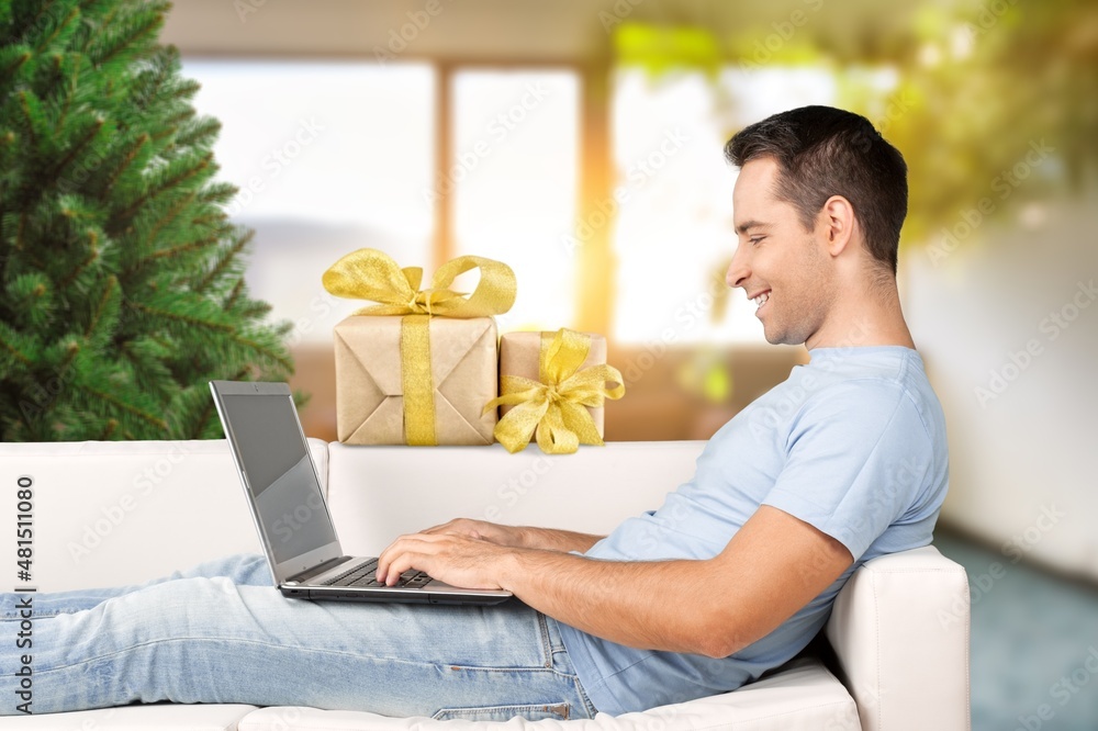 Playful man smiling sitting on the couch shopping online, buying Christmas presents