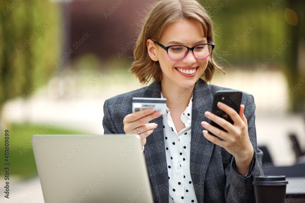 Positive business lady with mobile phone and credit bank card smiling at camera working remotely out