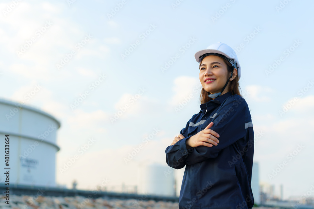 亚洲女工程师双臂交叉，面带微笑，自信地展望炼油厂的未来