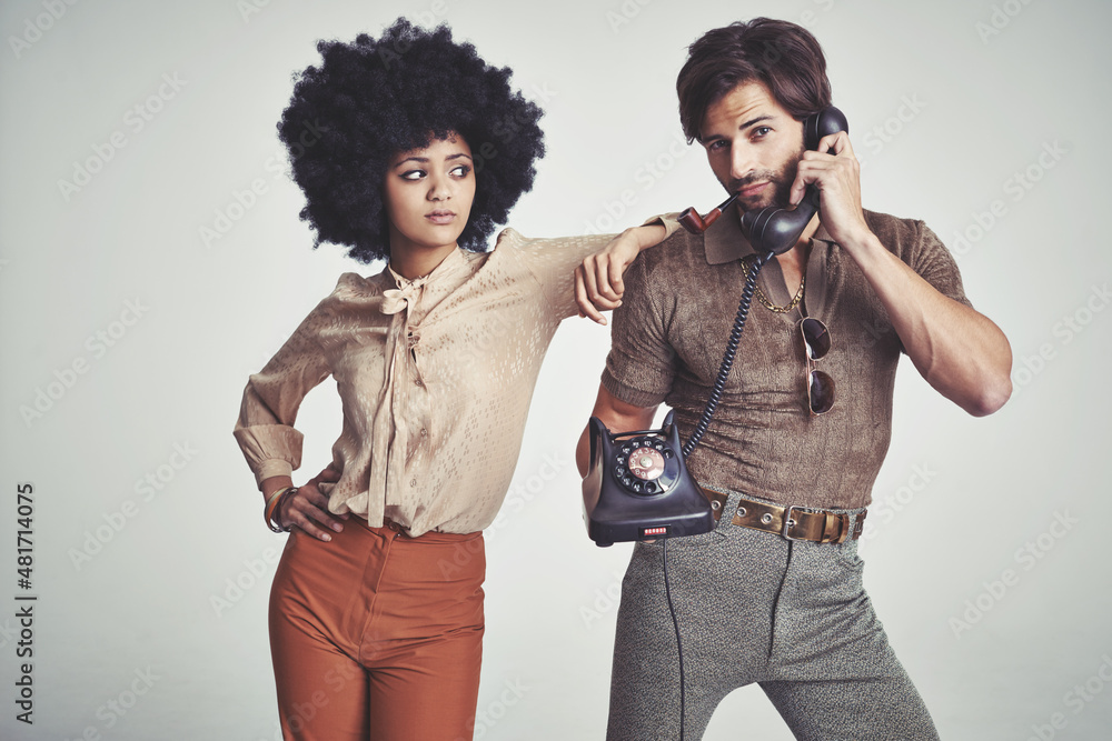 Taking that call. A beautiful young hippie looking over at a handsome man using a retro telephone.