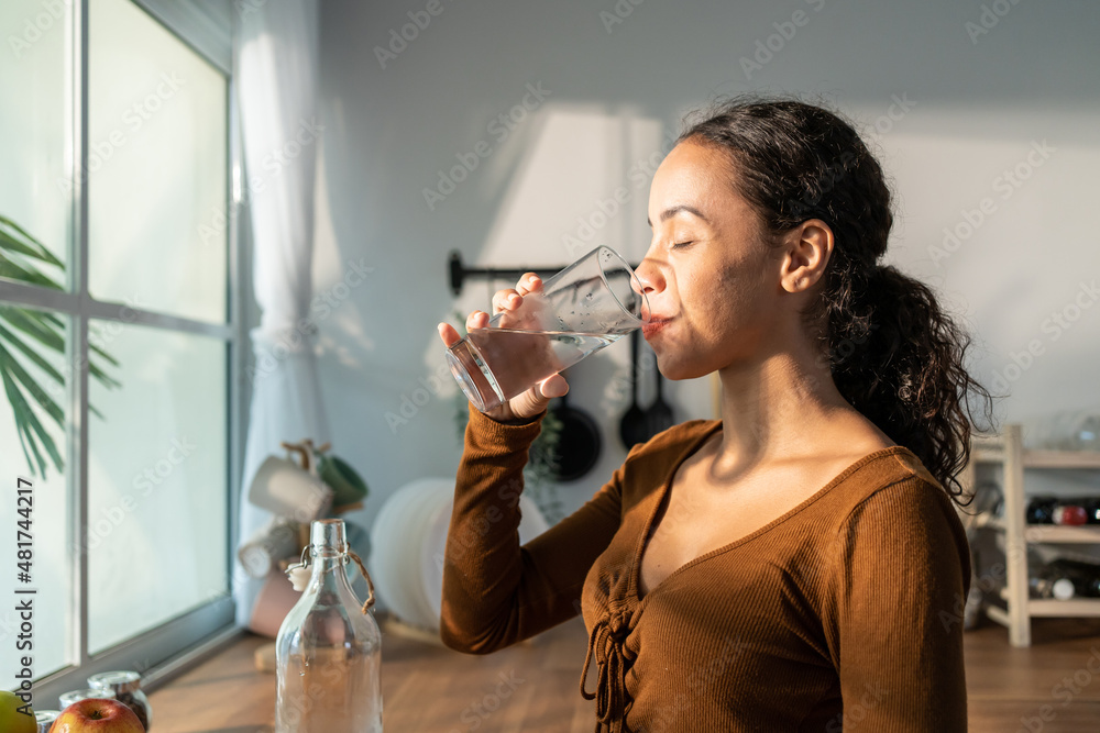 年轻漂亮的拉丁裔妇女在厨房里把干净的水放进玻璃里。