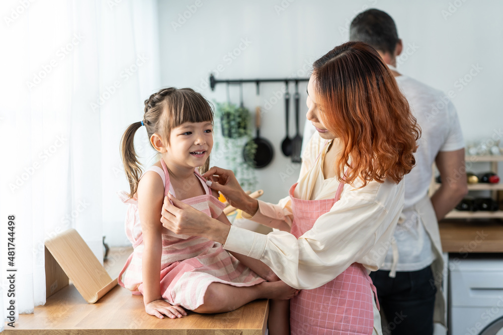 有魅力的亚洲父母在厨房里给可爱的小女儿穿围裙。