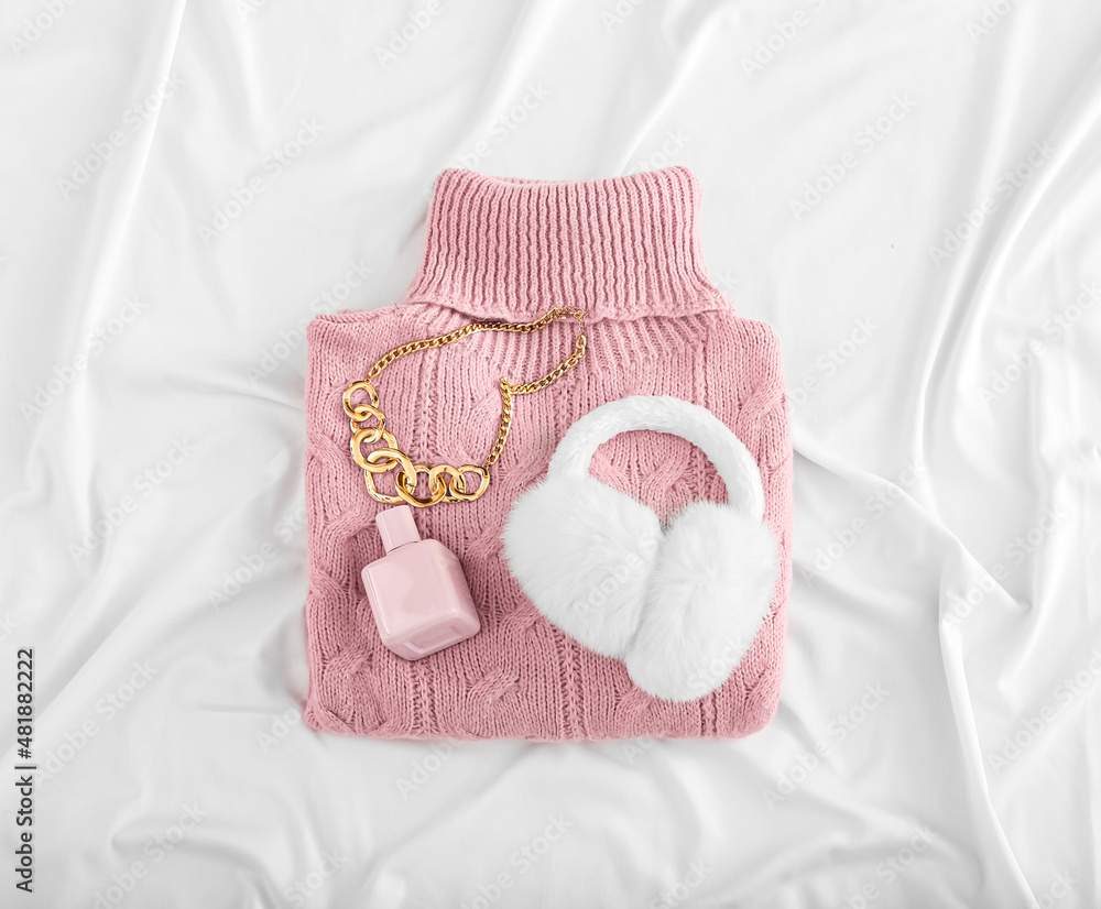 Knitted roll-neck sweater, perfume bottle and ear muffs on light crumpled fabric background