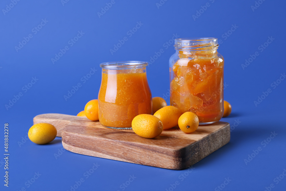 Jars of tasty kumquat jam and fresh fruits on blue background