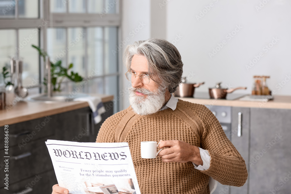 Senior man with cup of coffee reading newspaper at table in kitchen