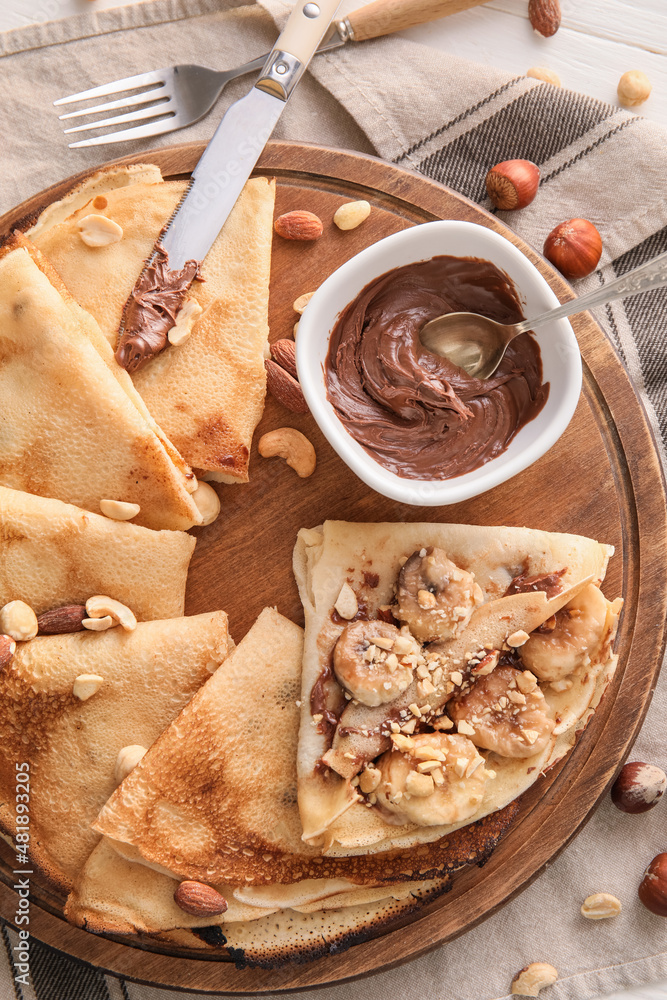 Board of tasty thin pancakes with chocolate paste, cut banana and nuts on table, closeup