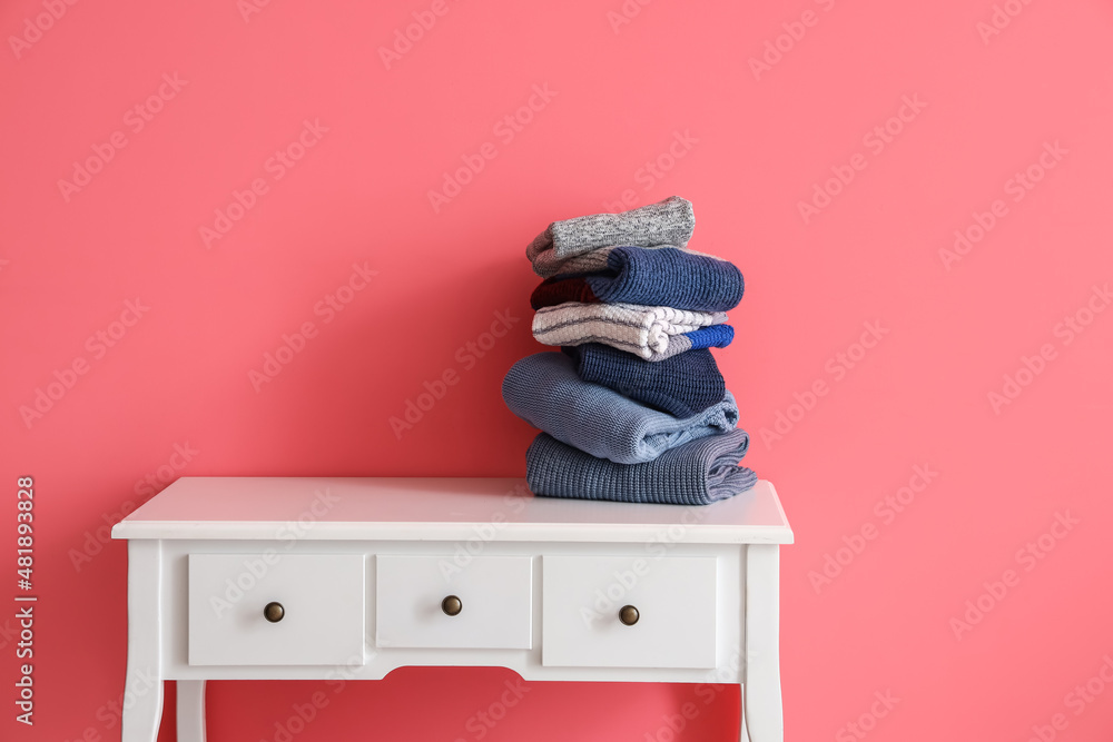 Stack of warm knitted sweaters on table near pink wall