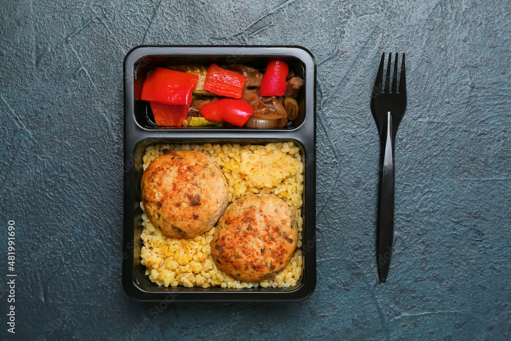 Food delivery container with delicious pearl barley, cutlets and vegetables on dark background