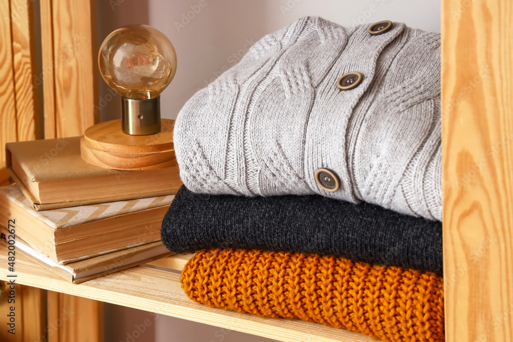 Stack of warm sweaters, books and lamp on shelf near light wall, closeup