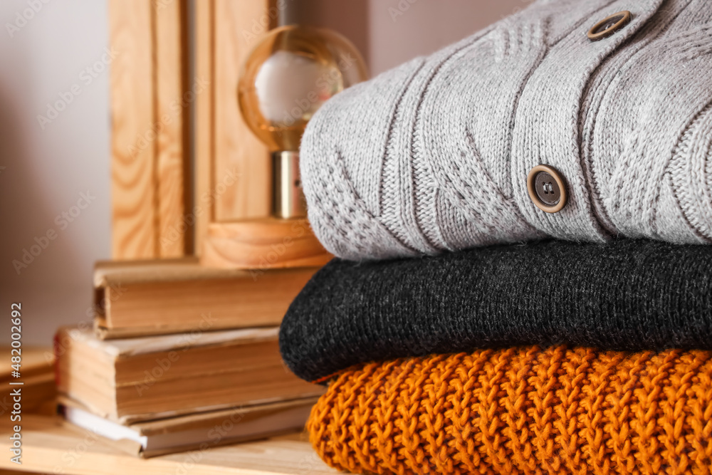 Stack of warm sweaters, books and lamp on shelf near light wall, closeup