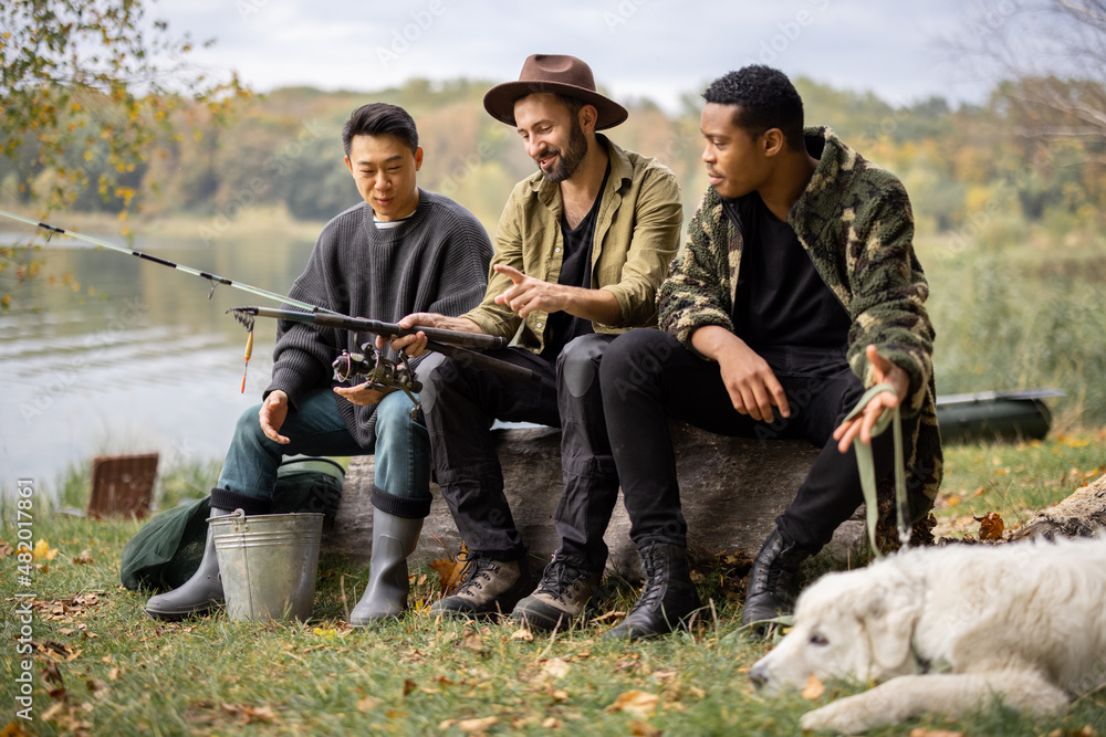 Multiracial male friends resting and talking in nature. Men after fishing on river or lake coast. Co