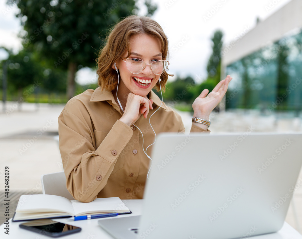 Happy young female teacher in earphones sitting in front of laptop outside having lesson online