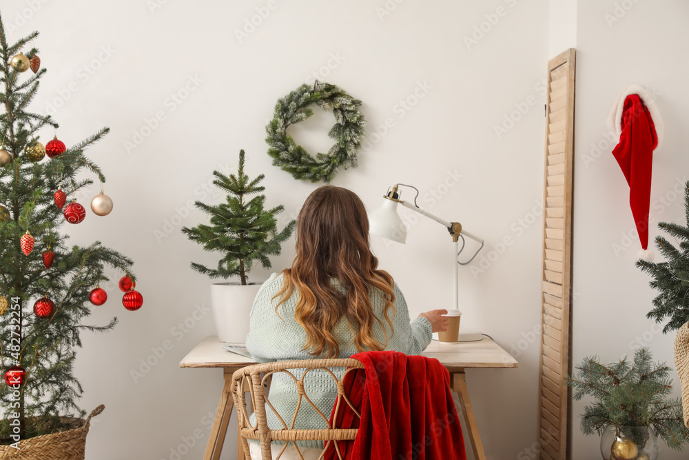 Woman sitting at table and taking paper cup with drink in room decorated for Christmas