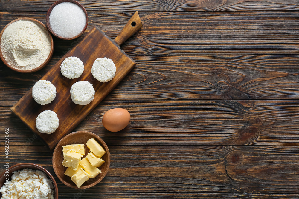 Board with uncooked cottage cheese pancakes and ingredients on wooden background