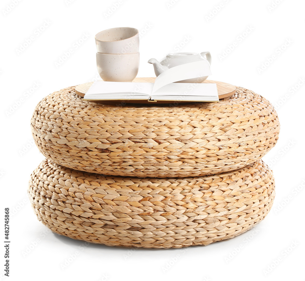 Opened book, cups and teapot on wicker poufs against white background