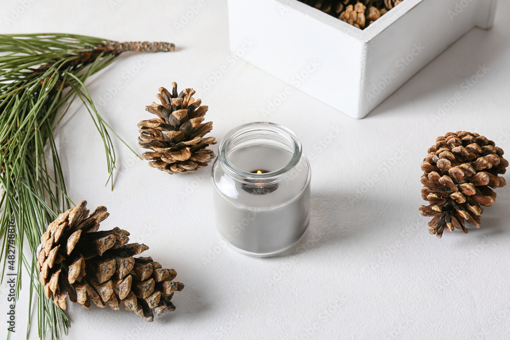 Glass holder with burning candle and pine cones on light background