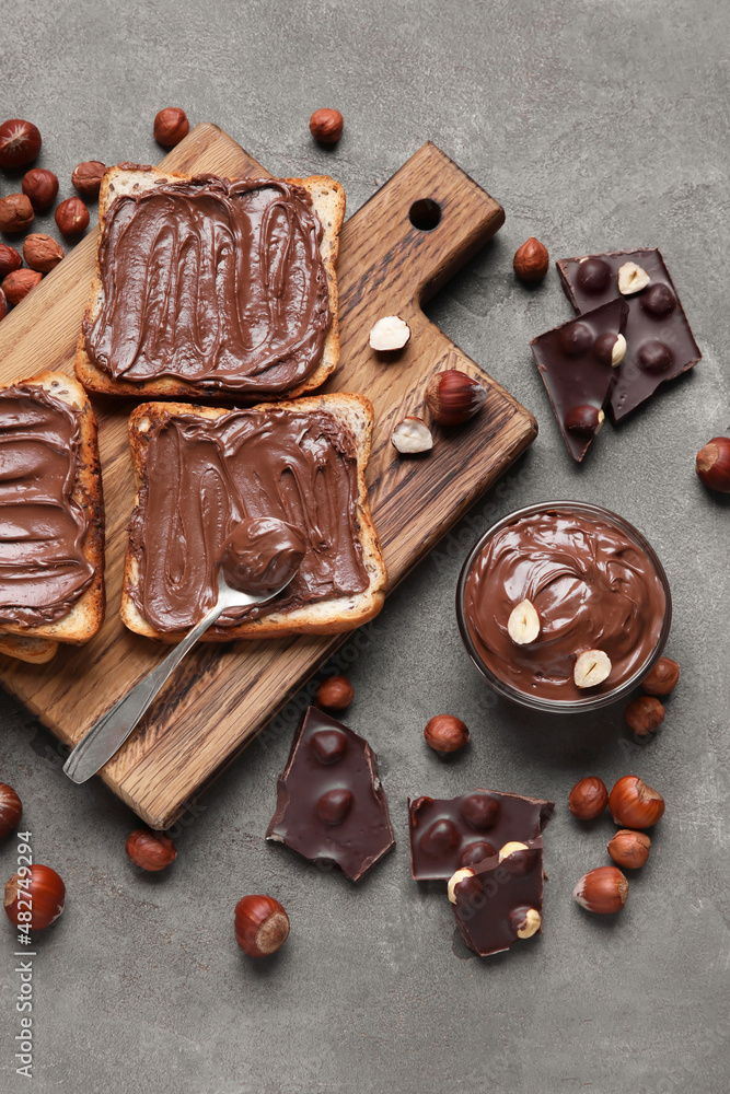 Wooden board of bread with chocolate paste and hazelnuts on grey background